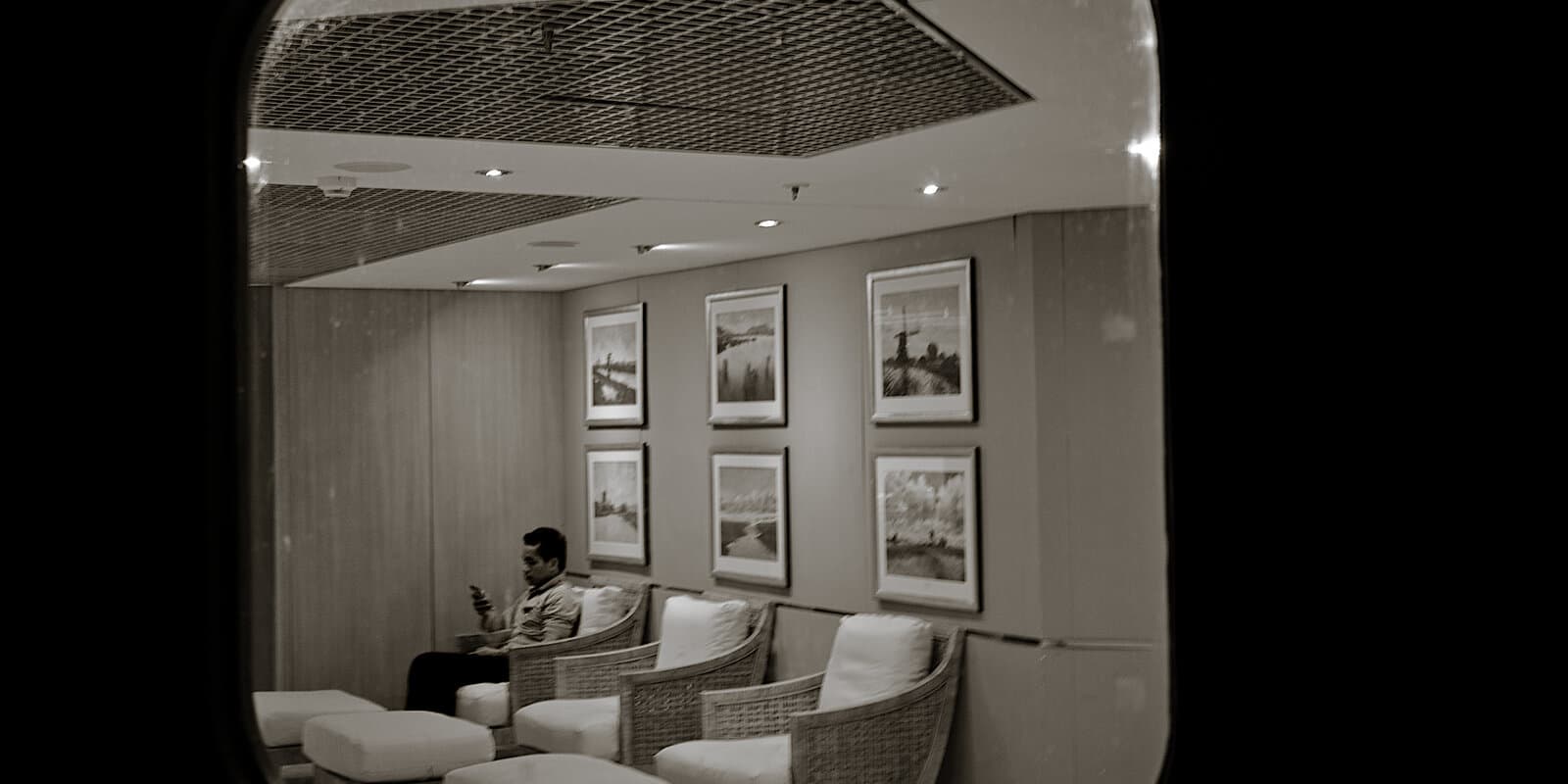 A person uses a smartphone in a quiet lounge on a cruise ship, seen through a deck door with wicker seating and framed artwork along the wall.