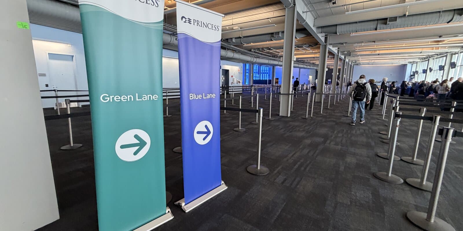 Travelers queue in lane lines inside a cruise terminal check-in hall with stanchions and directional banners